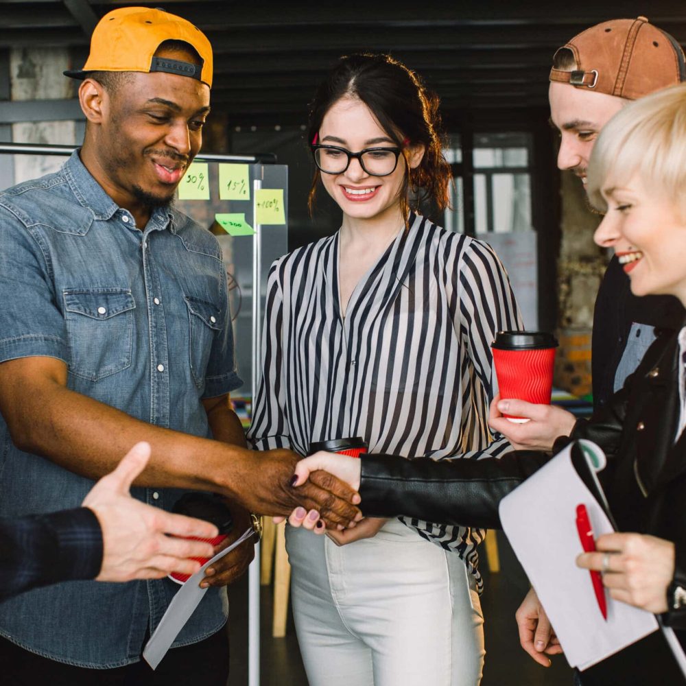 Diverse multiracial young people talking drinking coffee using devices in cozy office, multi-ethnic workers enjoy meeting.