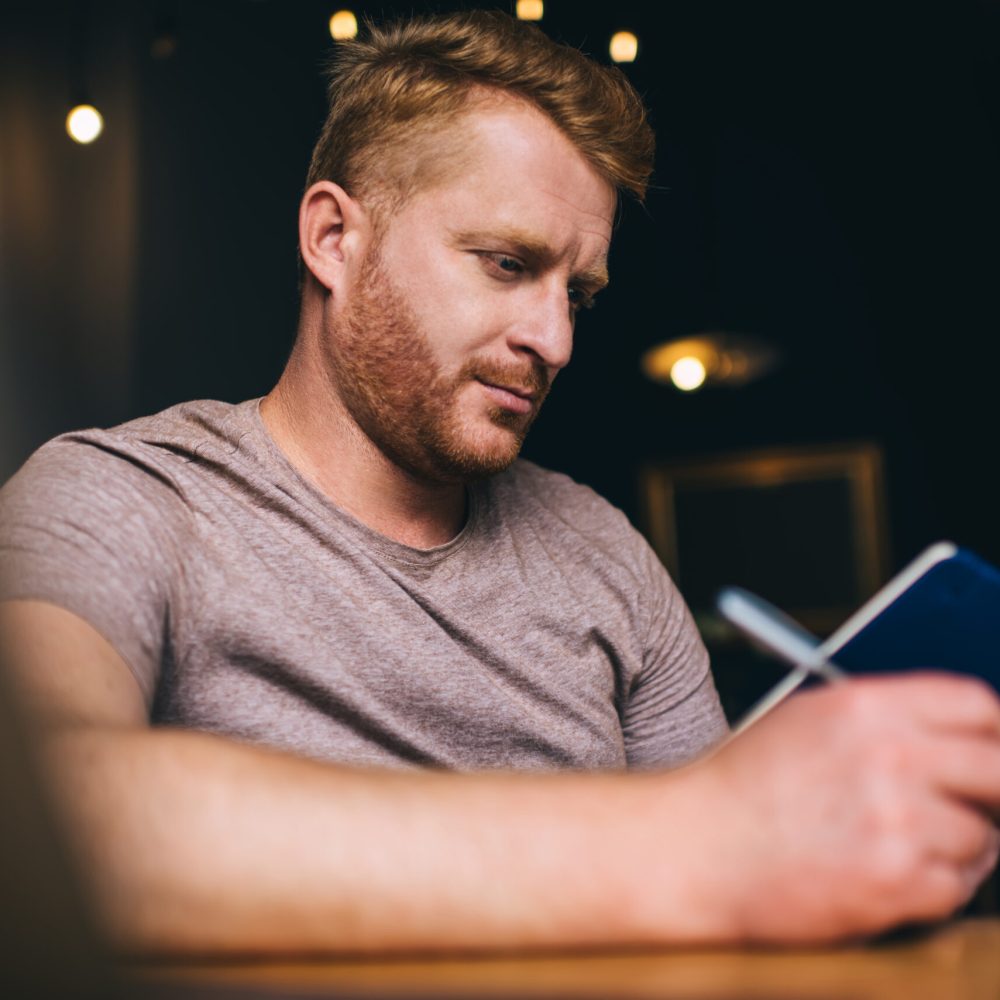 Concentrated male student sitting with notebook and pen and analyzing information while studying remotely from home with laptop during exam preparation